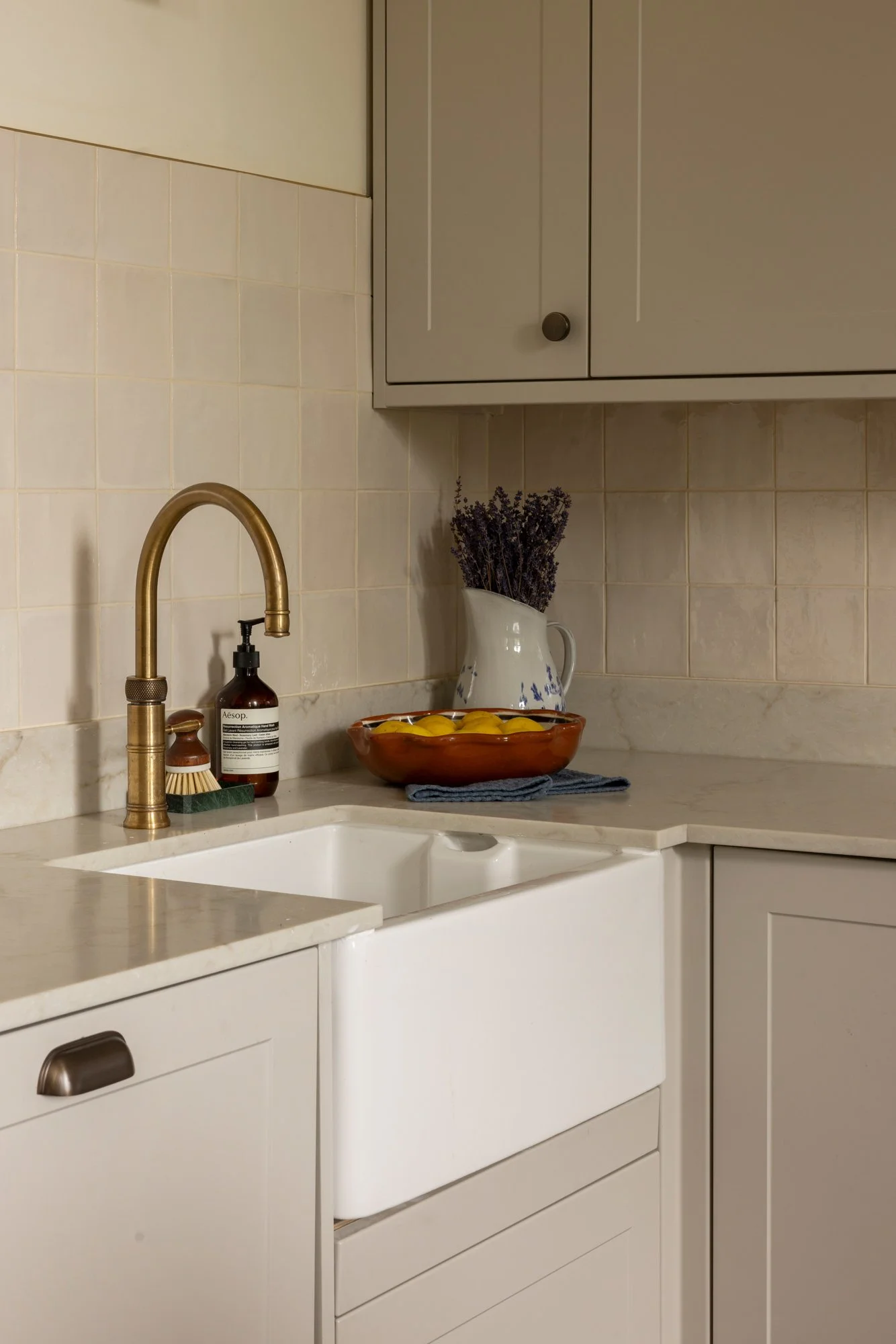 A kitchen countertop with a white farmhouse sink, a brass faucet, a brown soap dispenser, a brush, a white pitcher with lavender, and a bowl of lemons. White cabinets are above, with beige tiled walls.