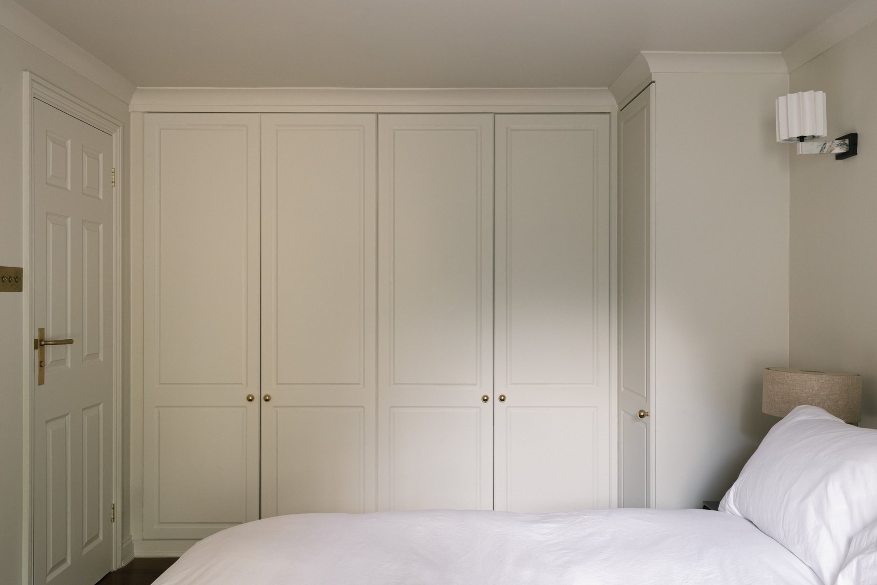 A bedroom with white walls, a large built-in white wardrobe, a bedside lamp with a beige shade, and part of a bed with white bedding.