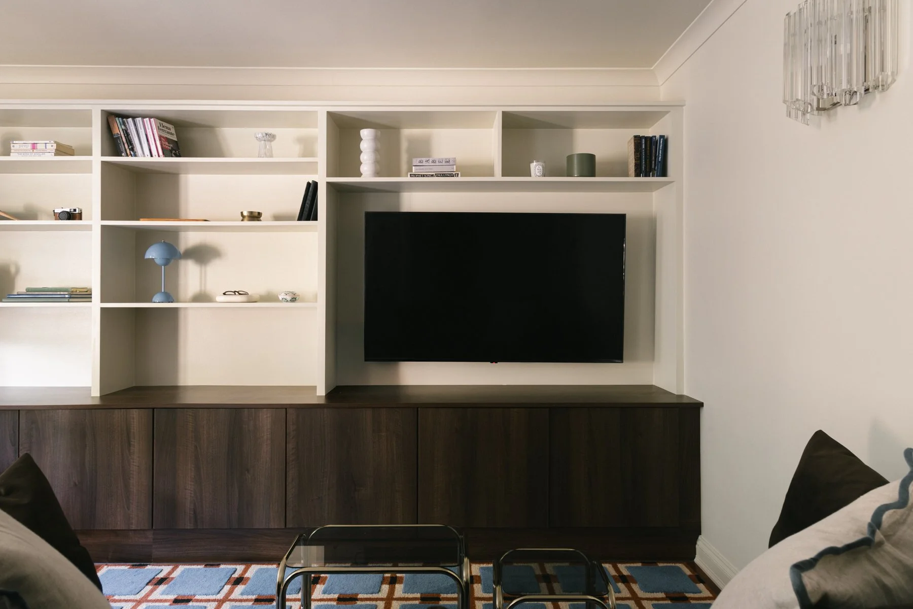 Living room with white built-in shelves, a flat-screen TV, dark wooden cabinet, and a patterned area rug. The shelves hold books, decorative objects, and a small camera. Part of a gray sofa with a throw pillow and a glass coffee table are visible in the foreground.