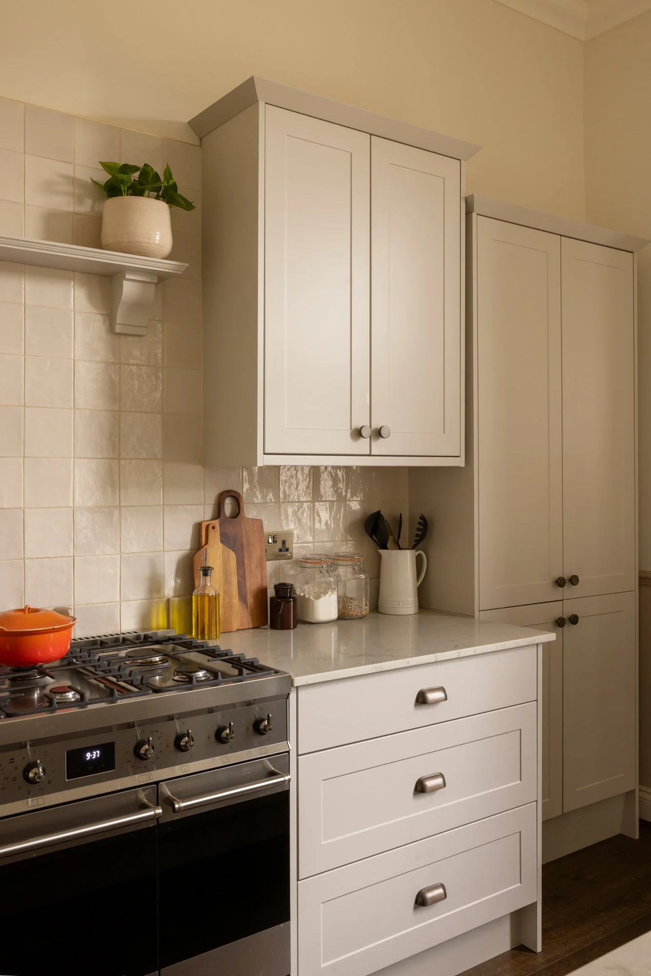 A kitchen with white cabinets, a white countertop, a stove with pots, and a tiled wall. Shelves hold a potted plant and various kitchen items.
