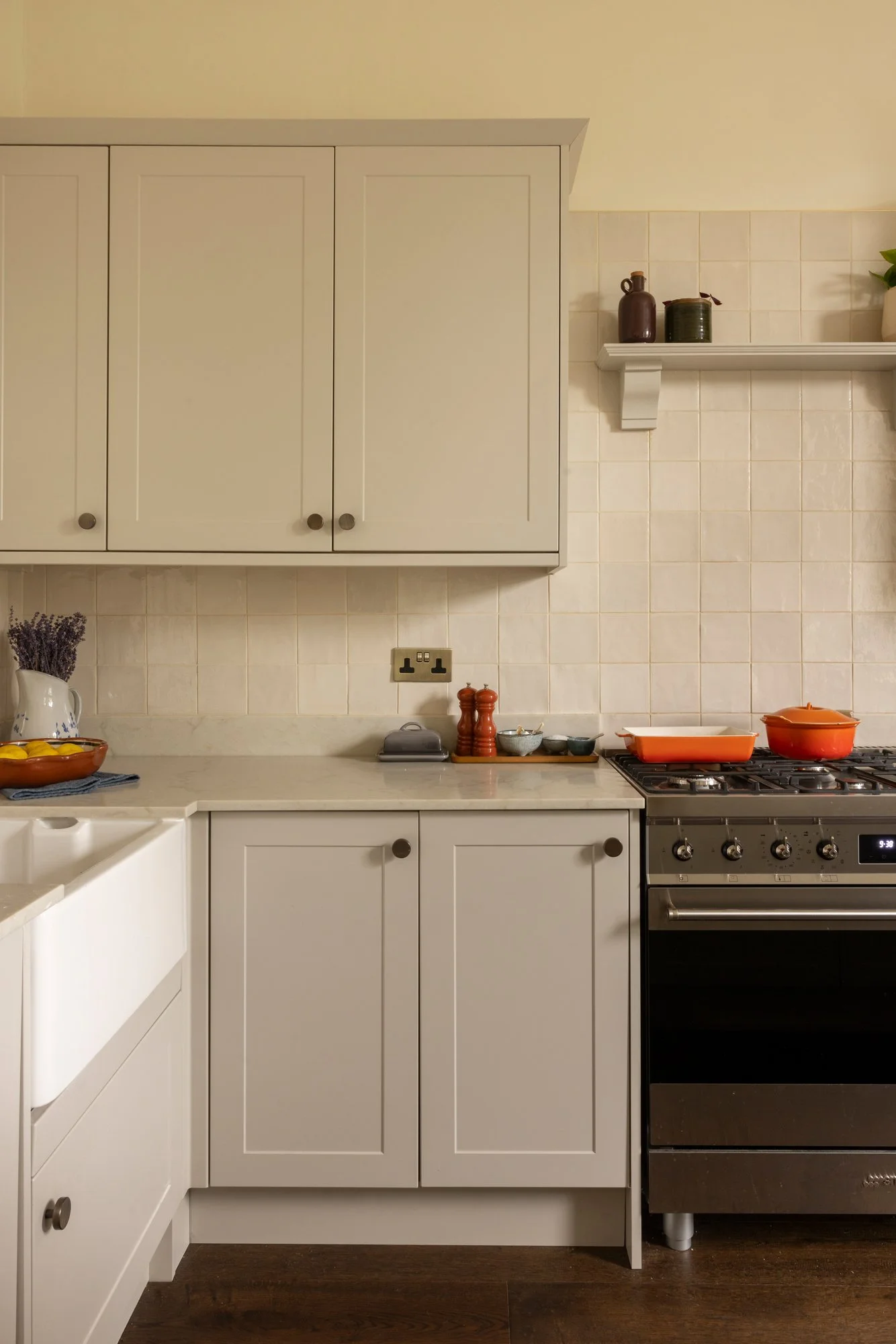 A kitchen with white cabinets, a gas stove with oven, and a white countertop. The backsplash is tiled in light cream. There are small decorative items on the shelf, and kitchen utensils and containers on the counter.