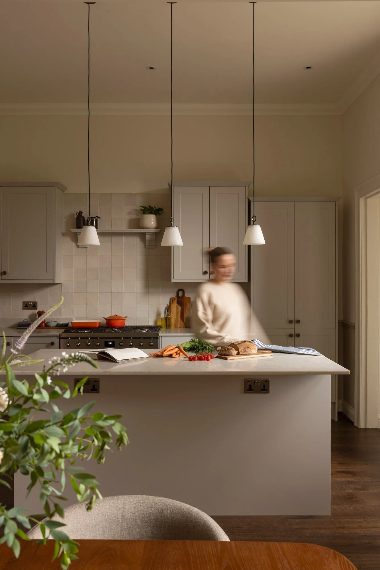 Kitchen scene with white cabinets, hanging pendant lights, a blurred woman in a beige top preparing food at a white kitchen island with vegetables and bread, and a plant in the foreground.