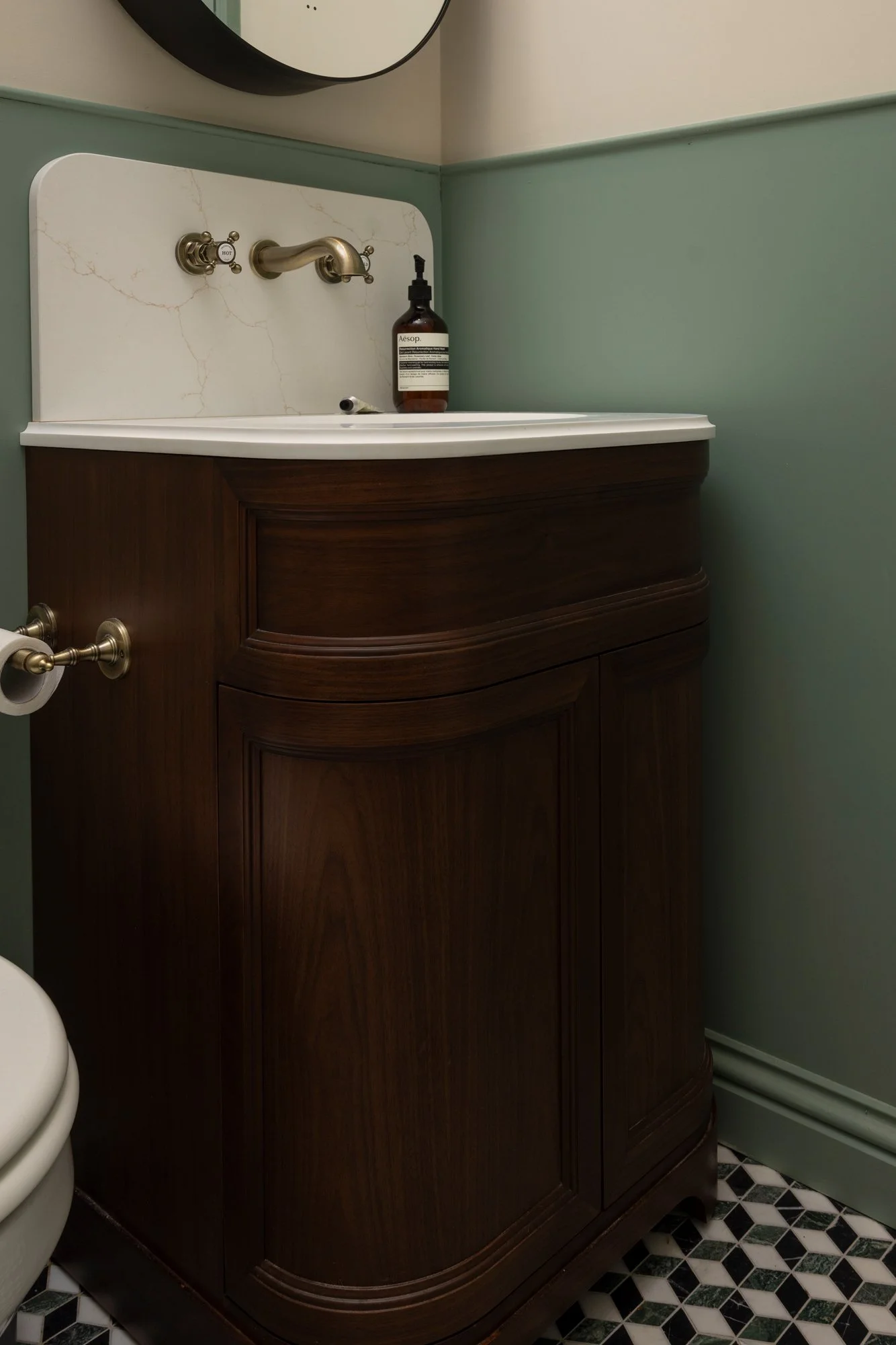 Bathroom sink with brass faucet, bottle of hand soap, mirror with black frame, wooden cabinet, and patterned floor tiles.