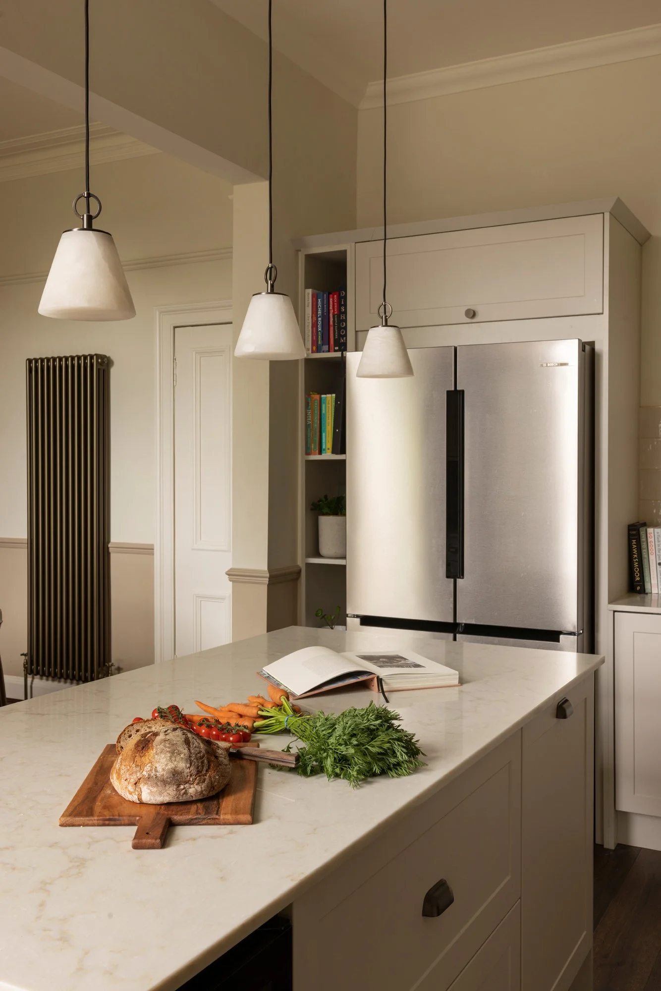A kitchen with a white marble island counter, fresh vegetables including carrots, tomatoes, basil, and bread on a cutting board, a book open on the counter, a refrigerator, bookshelves, and hanging pendant lights.