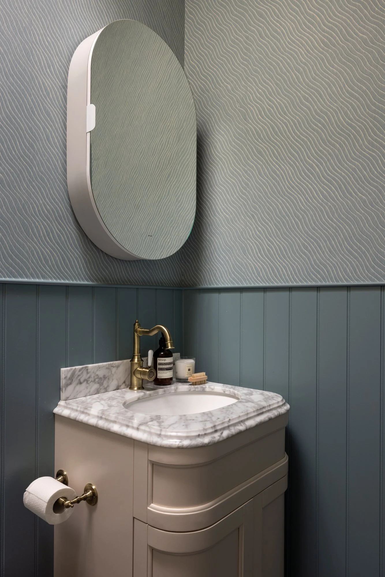 Bathroom vanity with marble countertop, oval mirror, gold faucet, toiletries, and a blue wavy patterned wall.