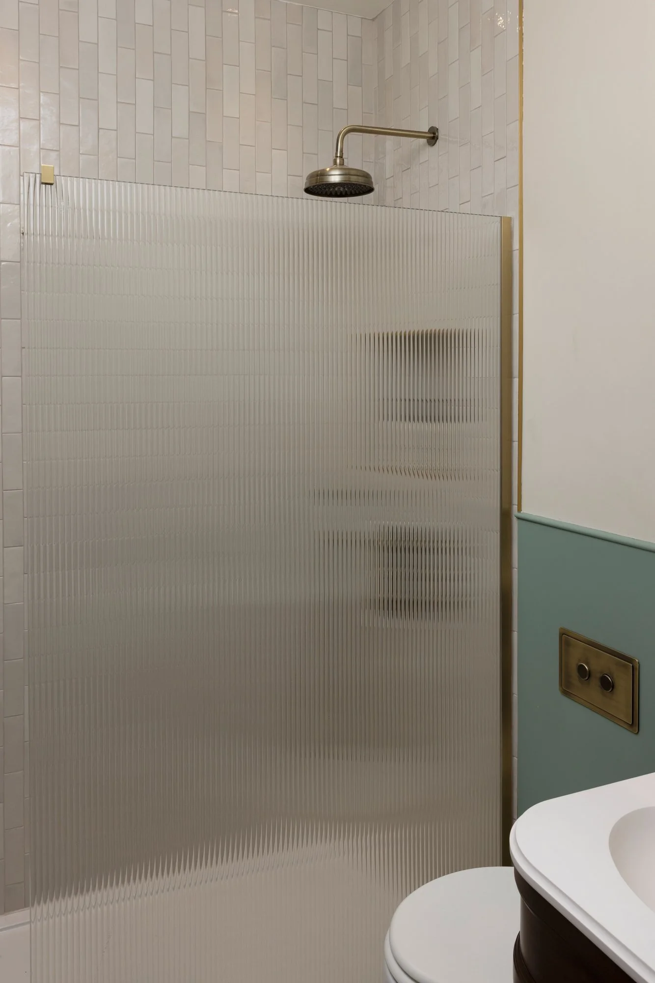 A restroom with a shower area enclosed by a frosted glass partition. The shower has white tiled walls and a wall-mounted shower head. To the right, there are two toilet bowls side by side. The wall beside the toilets has an electrical outlet.