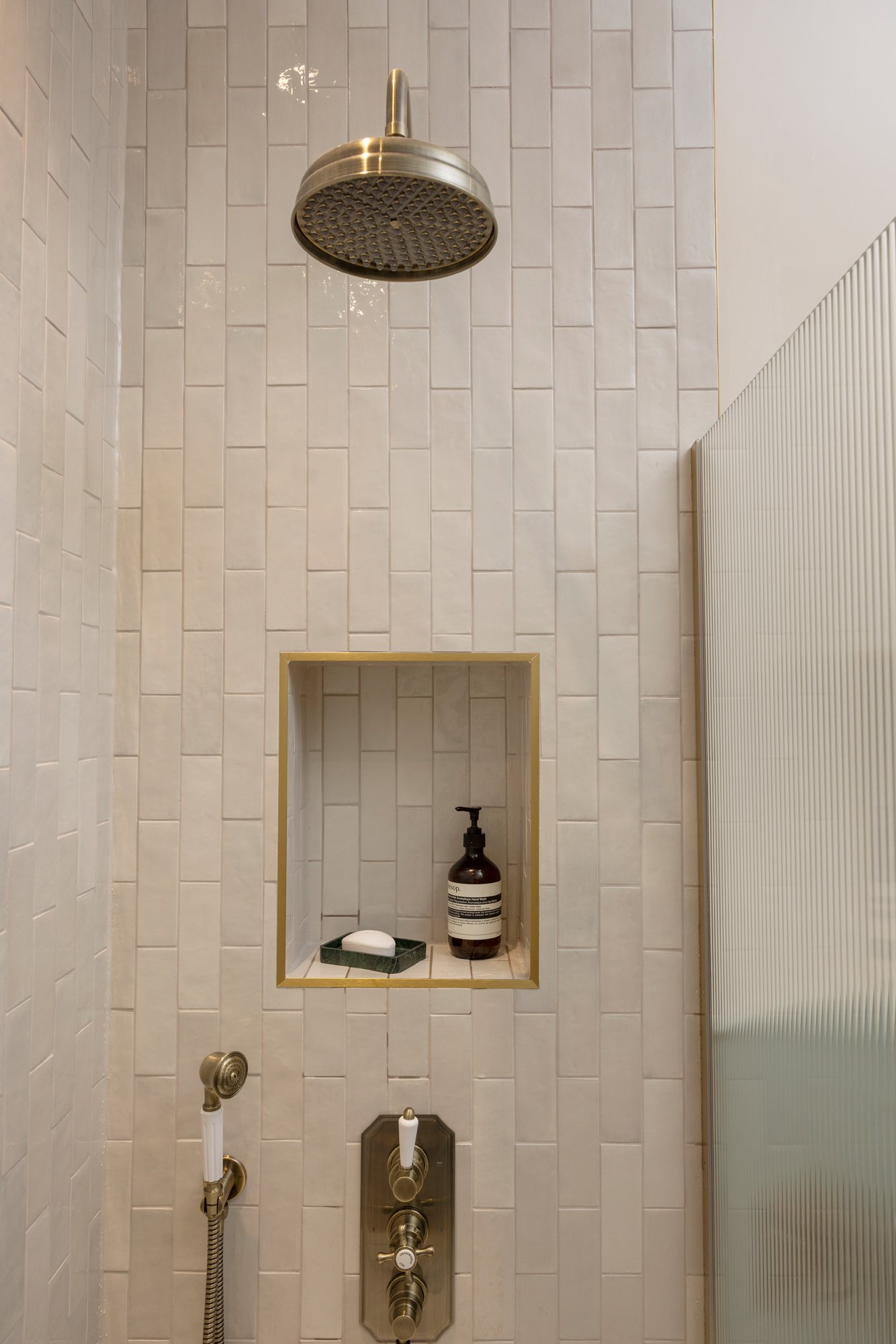 Shower area with a large showerhead, a built-in niche with a bottle and soap, and multiple controls for water, set in white rectangular tiled wall.