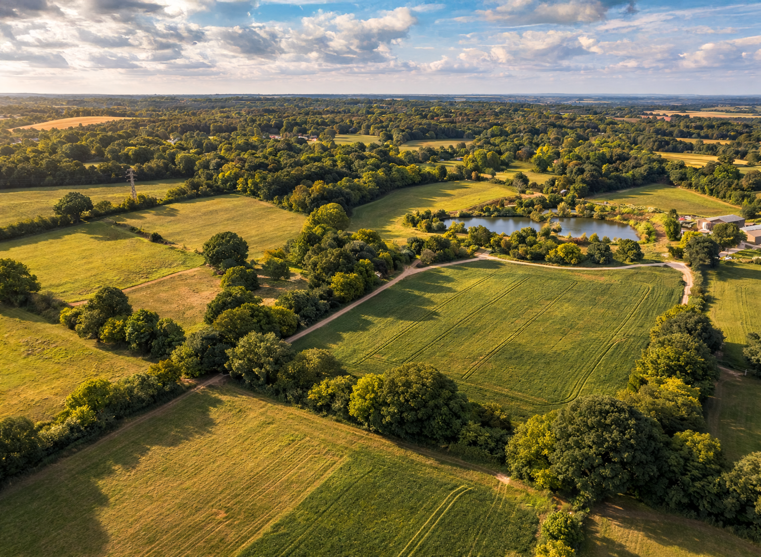 Aerial view of rural landscape with green fields, trees, a small lake, and dirt roads under a partly cloudy sky.
