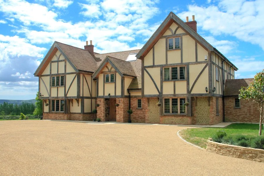Large Tudor-style house with brick and half-timbered exterior, multiple gabled roofs, and a spacious front yard under a partly cloudy sky.