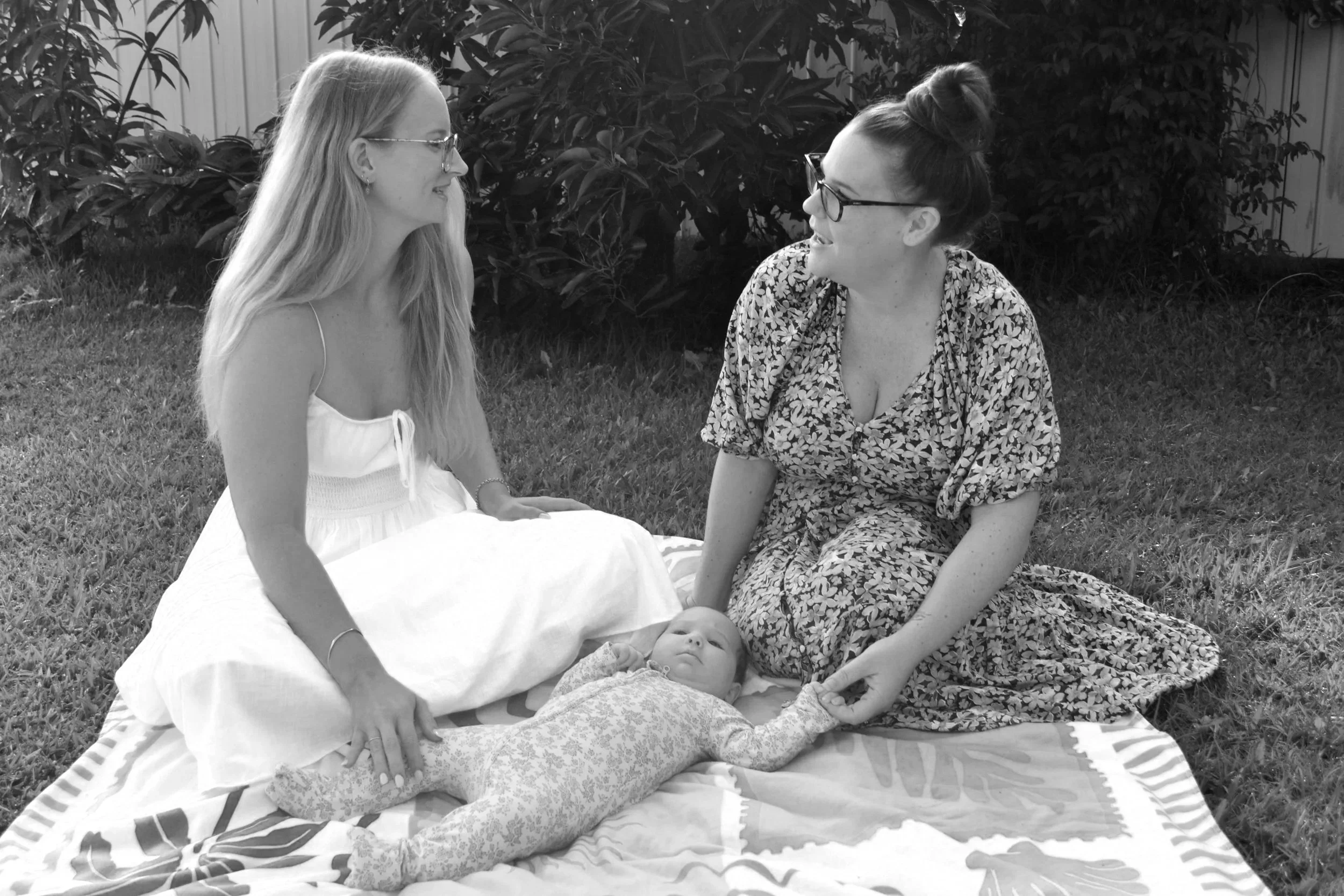 Two women sitting on grass outside, one with long hair and glasses wearing a white dress, the other with a bun and glasses wearing a patterned dress, with a baby lying on a blanket in front of them.