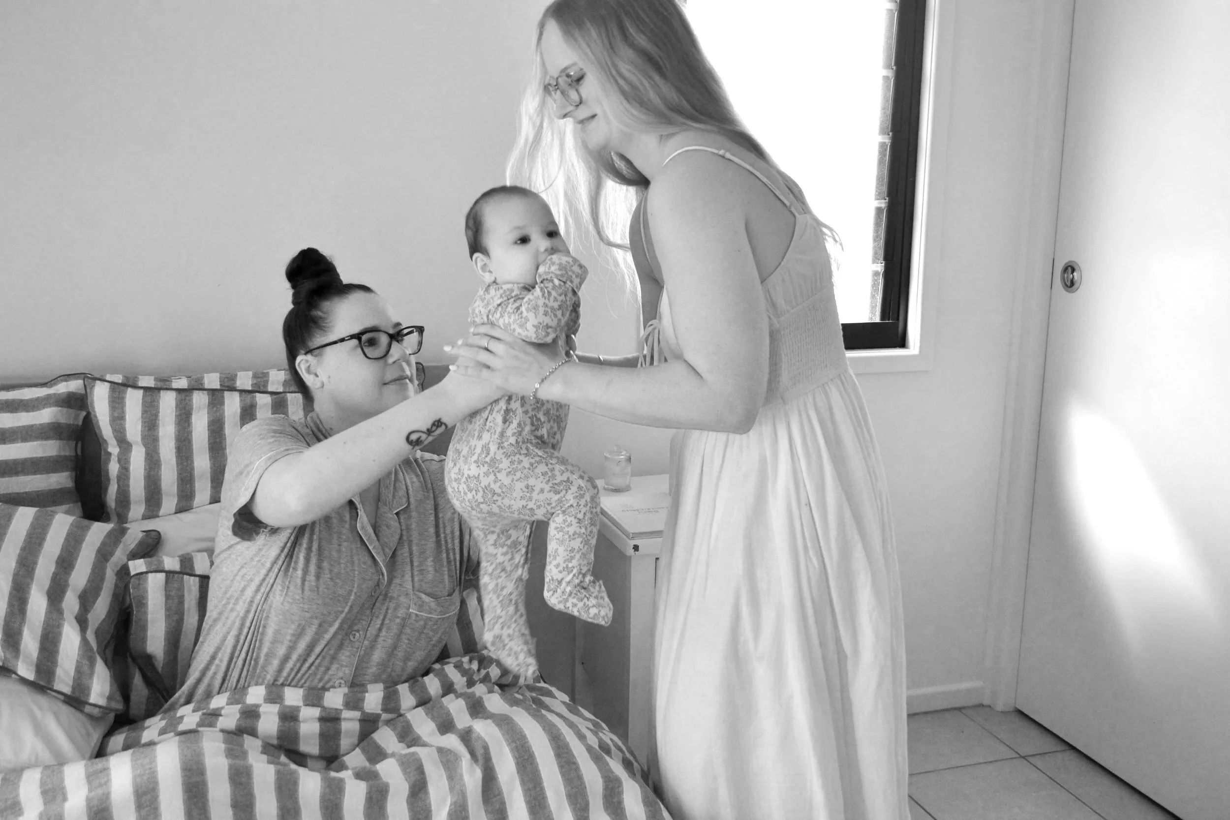 A woman sitting on a bed with striped pillows holds a baby while another woman in a white dress leans over them, appearing to interact positively.