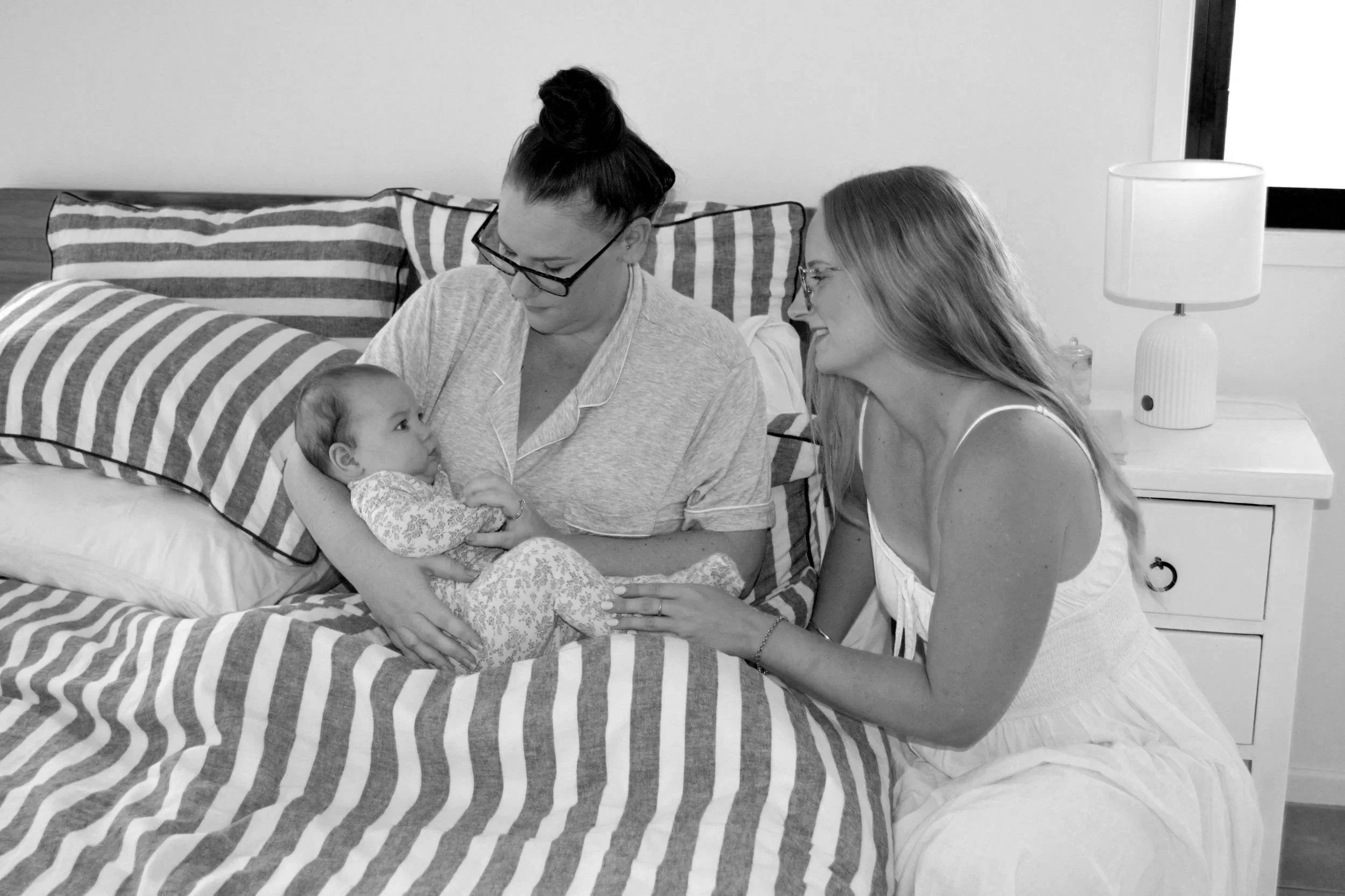 Two women sitting on a bed with a young girl, looking at her affectionately, in a bedroom with striped bedding and a nightstand with a lamp.