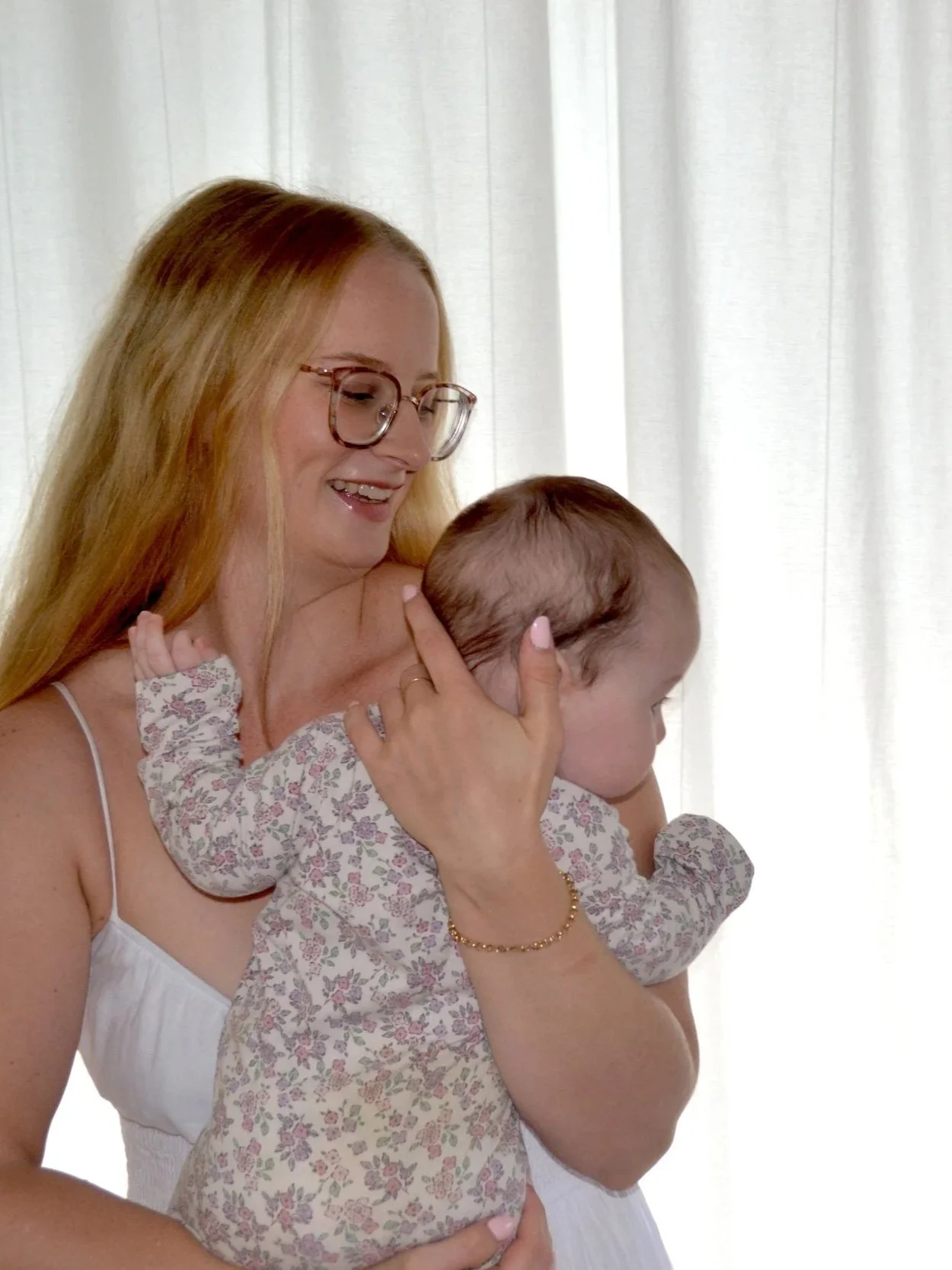 A woman with long red hair and glasses holding a young child with brown hair, both near a white curtain.