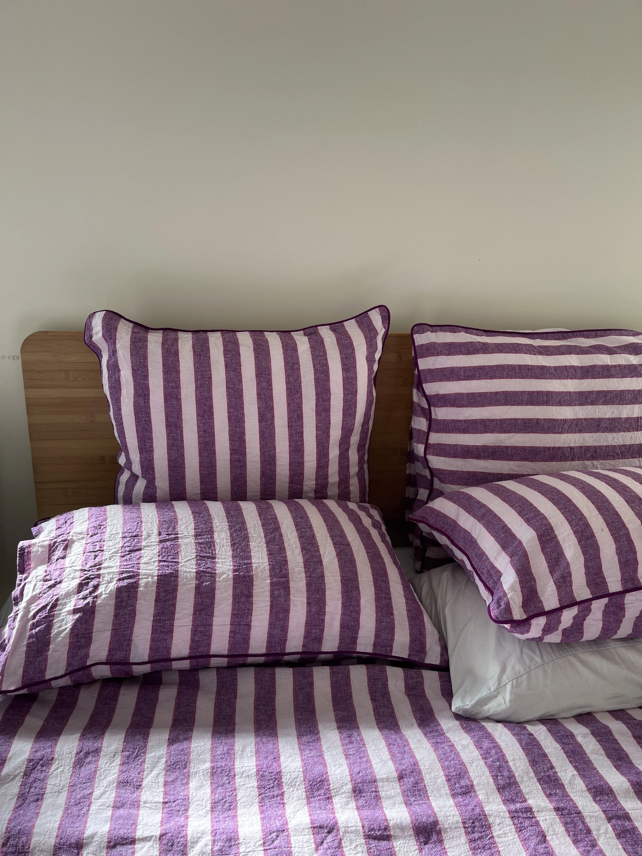 A bed with a wooden headboard, covered with pink and purple striped bedspread, and two matching pillows. One pillow is on the left and the other on the right, with a white pillow partially visible underneath on the right. The wall behind the bed is plain and light-colored.