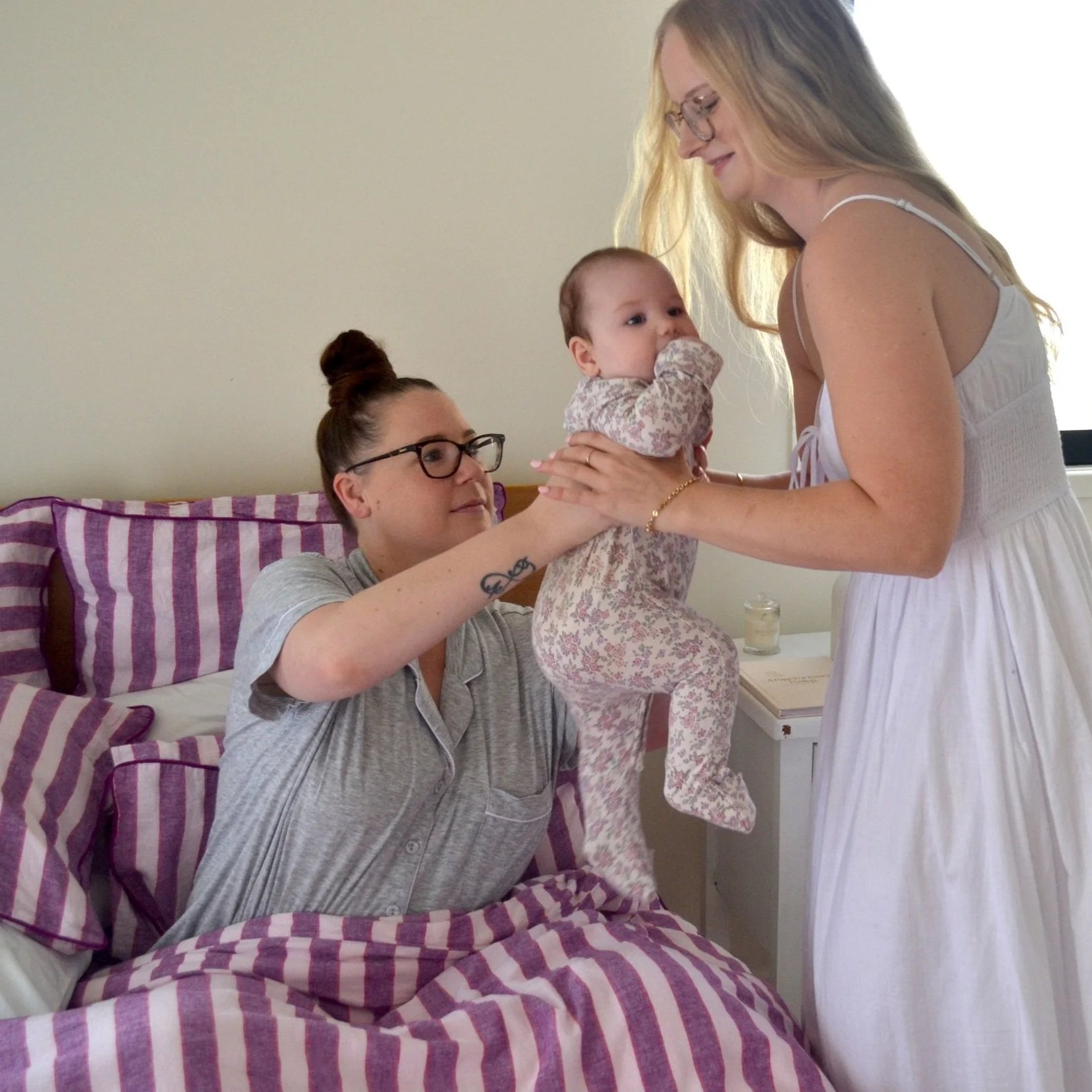 A woman in glasses and gray pajamas is sitting in bed, holding a young girl in floral pajamas with a pacifier in her mouth, while a woman in a white dress stands in front of them, all smiling and interacting warmly.