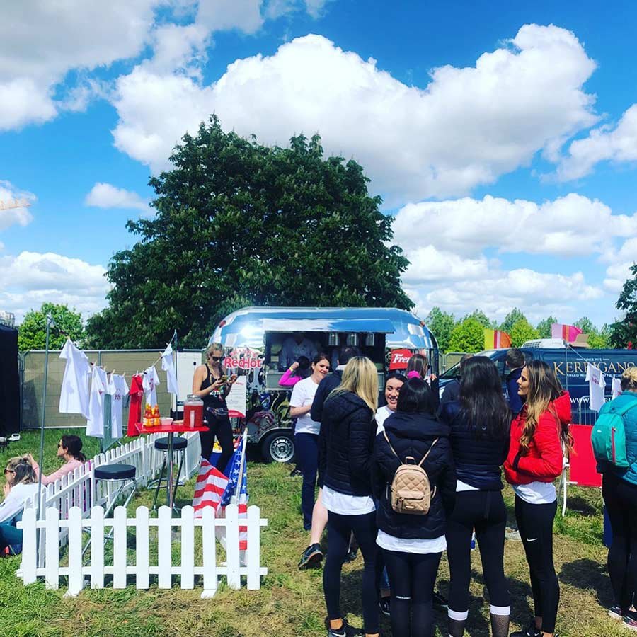 People gathered around a red, white, and blue food truck at an outdoor event on a partly cloudy day.