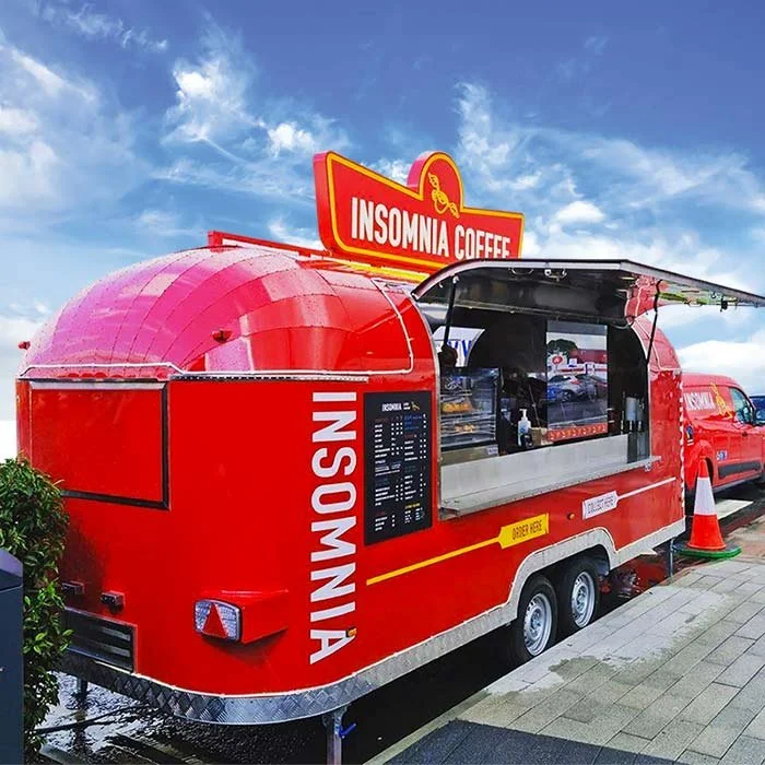 Red coffee food truck with 'Insomnia Coffee' signage, parked on sidewalk under a blue sky with clouds.