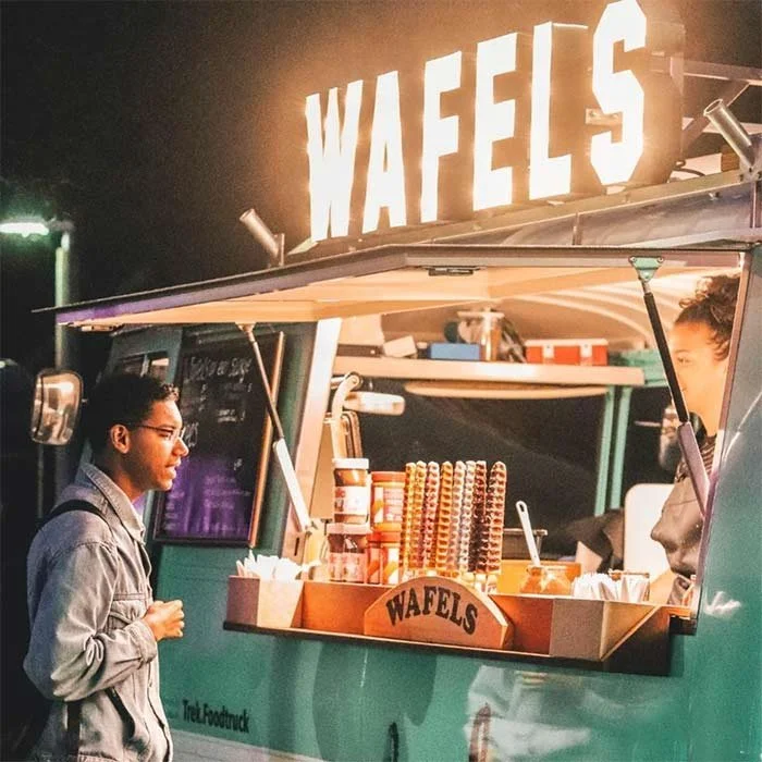 A food truck with a prominent sign reading "WAFELS" serving waffles, with a woman customer in front in a denim jacket and glasses.