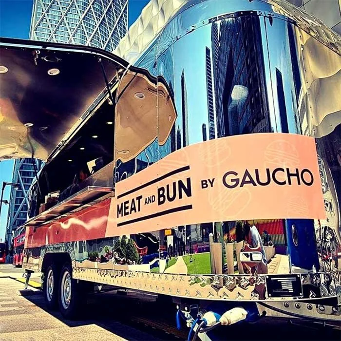 Food truck with a sign that reads "Meat and Bun by Gaucho," parked on a city street with tall buildings reflected in its shiny metallic surface.