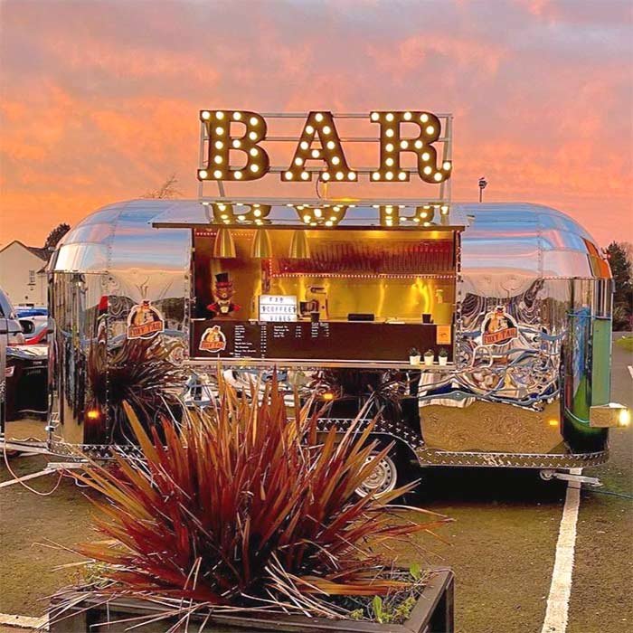 A shiny, silver, vintage-looking food trailer with a large illuminated "BAR" sign on top, set against a colorful sunset sky. Potted plants are in the foreground.