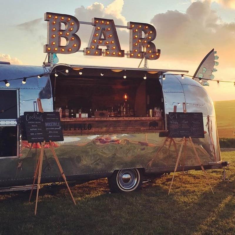 A vintage Airstream trailer converted into a bar, with a large illuminated sign spelling 'BAR' on top, set outdoors during sunset.