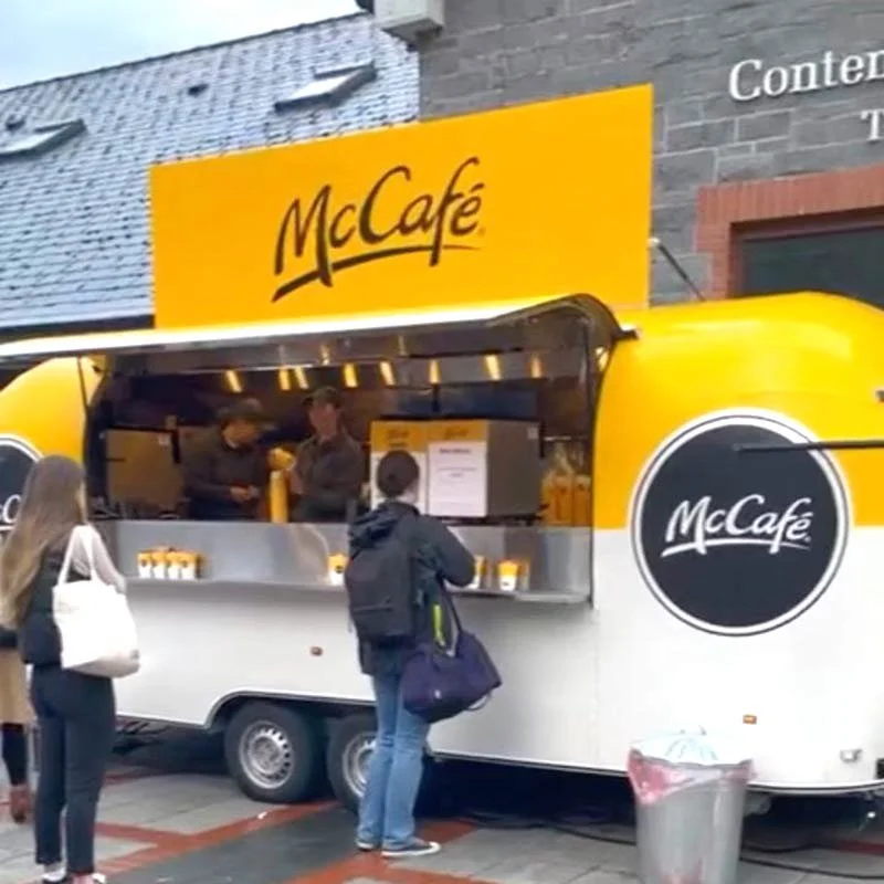 A McCafé food truck with a yellow and white exterior serving customers, with a group of people waiting to order.