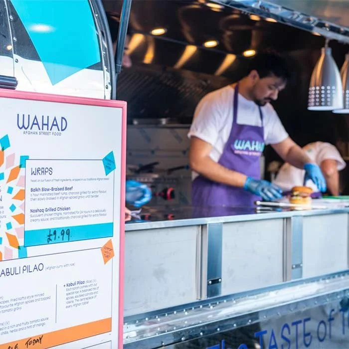 A man wearing a white shirt, purple apron, and blue gloves preparing food at a food truck named 'WAHAD'. A menu board displaying wraps, Balkh slow-brisket beef, and Noshak grilled chicken. The food truck has a stainless steel counter and hanging lamps.