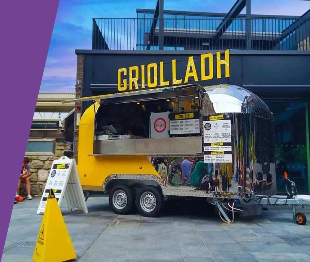A yellow food trailer with a shiny, reflective exterior parked outside a building with a sign reading 'Griolladh' at sunset.