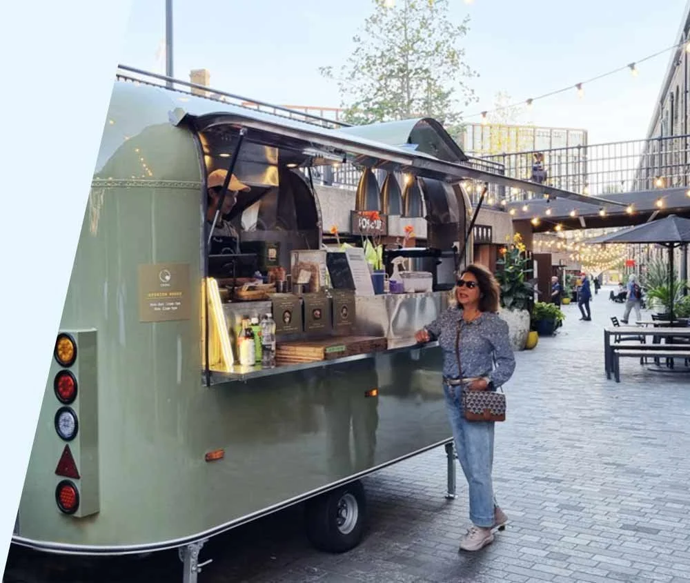 A woman standing outside a food truck on a city street, smiling and wearing sunglasses.