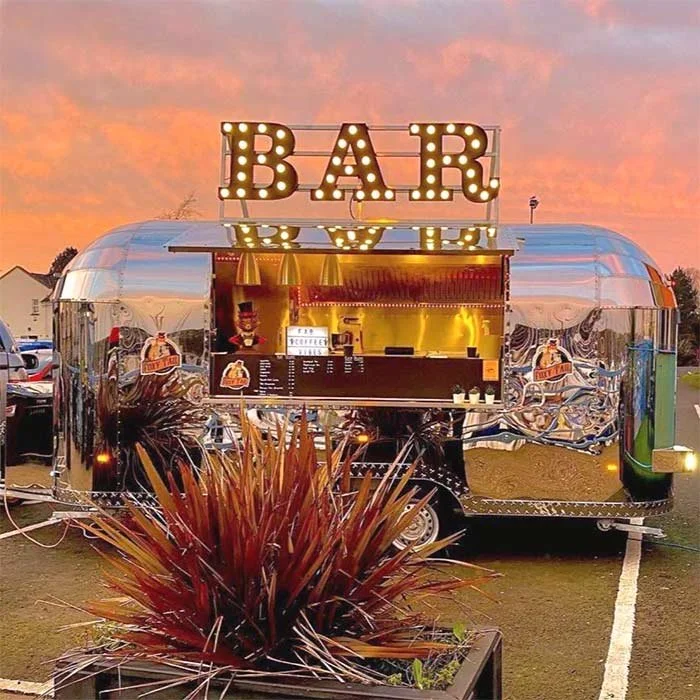A shiny silver Airstream trailer serving food and drinks as a bar with a large illuminated sign reading 'BAR' on top, set against a sunset sky.