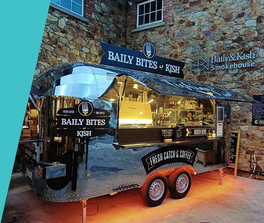 A mobile food stand named 'Baily Bites & Kish' serving fresh catch and coffee, parked in front of a stone wall with windows, with a sign on top and a small chalkboard on the side.