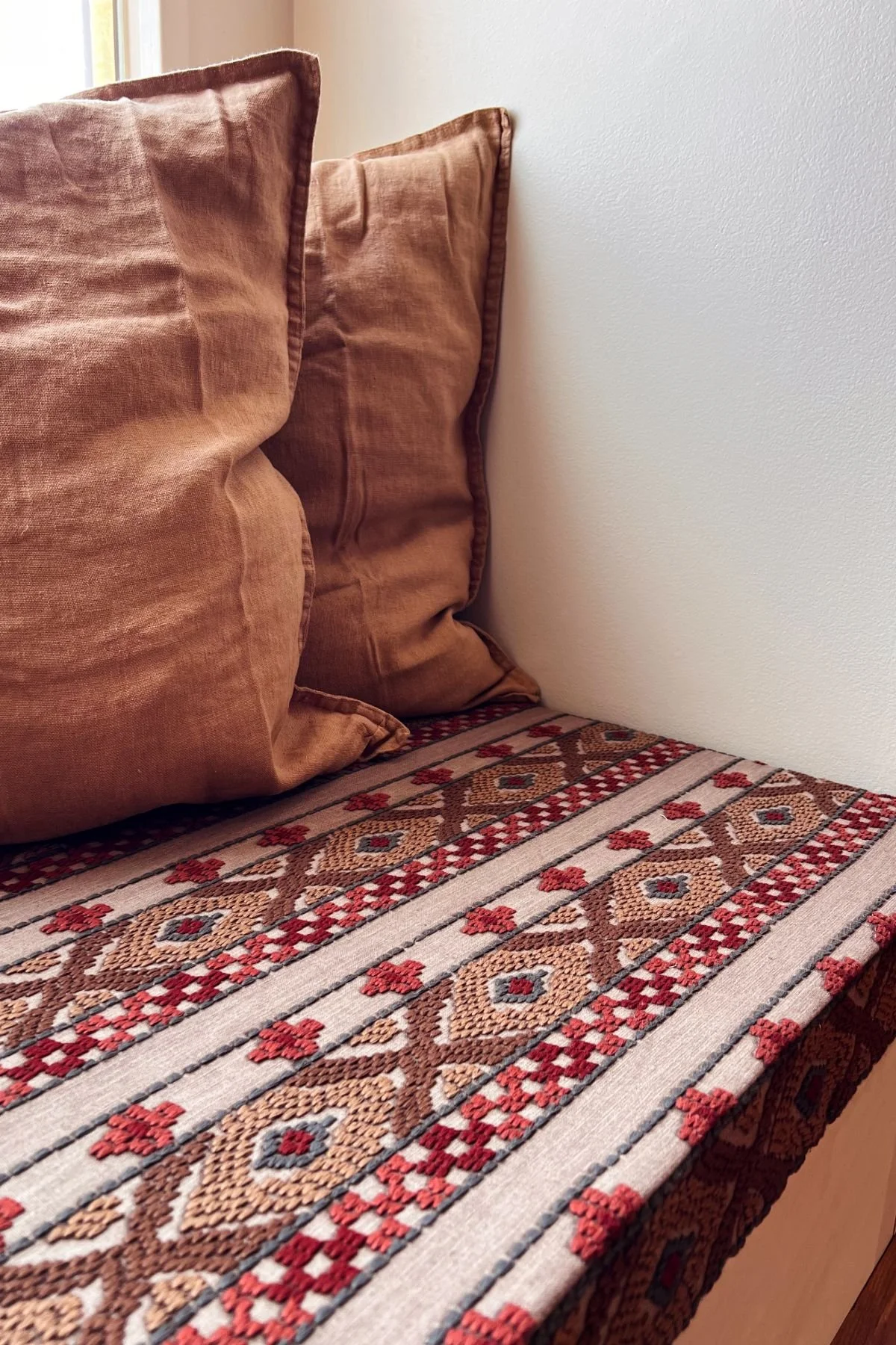 A bed with decorative pillows and a patterned bedspread featuring red, beige, and black geometric designs, against a white wall and near a window.