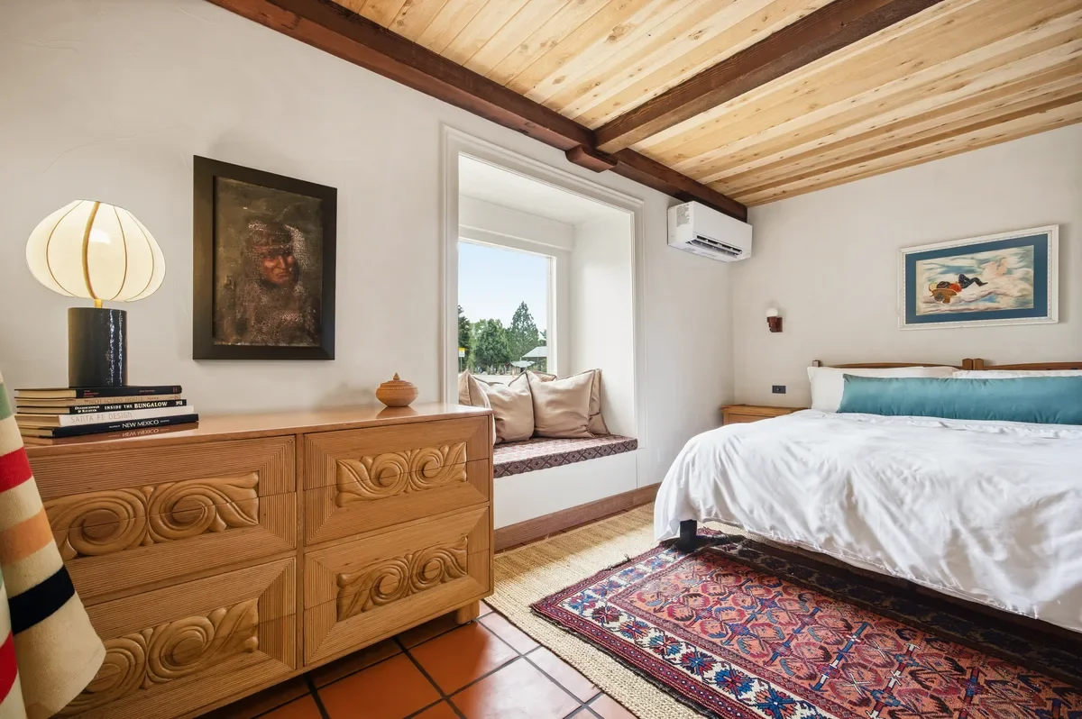 A cozy bedroom with a wooden ceiling, a large bed with white bedding and a long blue pillow, a window seat with cushions, a wooden dresser with books and a lamp, framed artworks on the walls, a colorful patterned rug, and an air conditioning unit.