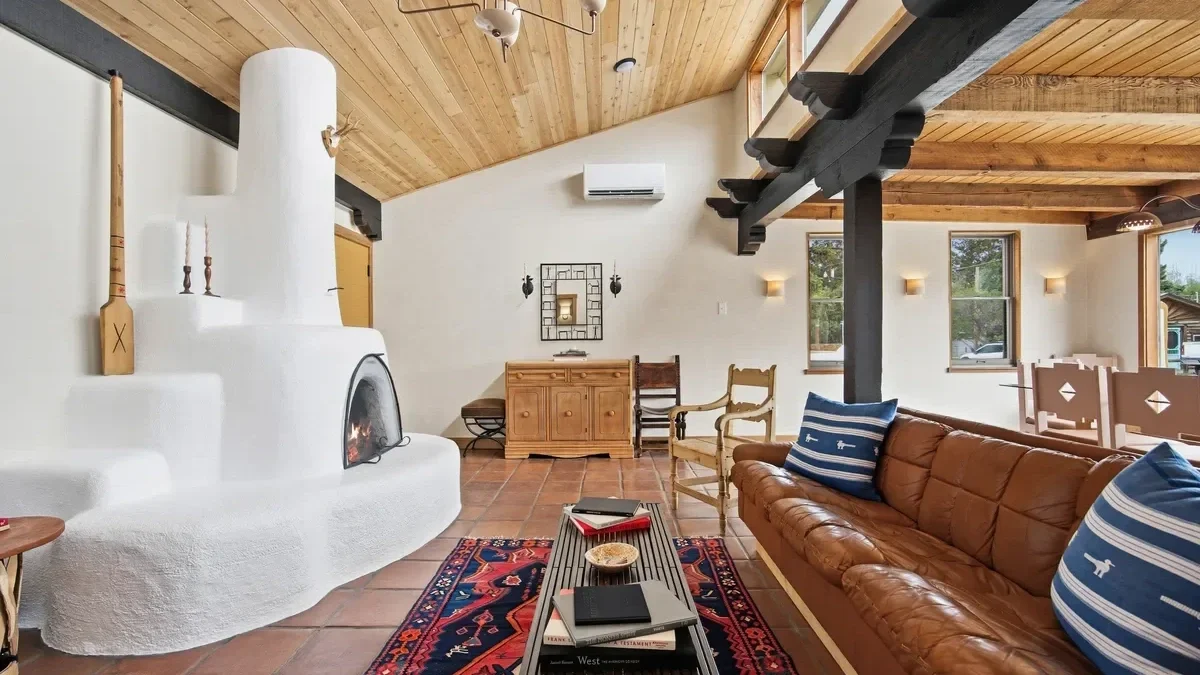 Living room with white stucco fireplace, brown leather sofa with striped pillows, wooden ceiling with beams, and rustic furniture