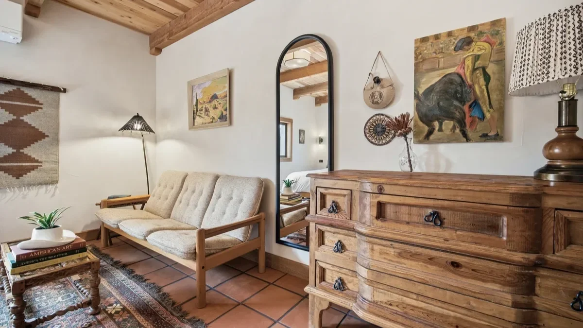 Living room with beige tufted sofa, wooden coffee table with books and plant, wall art, a large mirror, wooden dresser, and various decorative items.