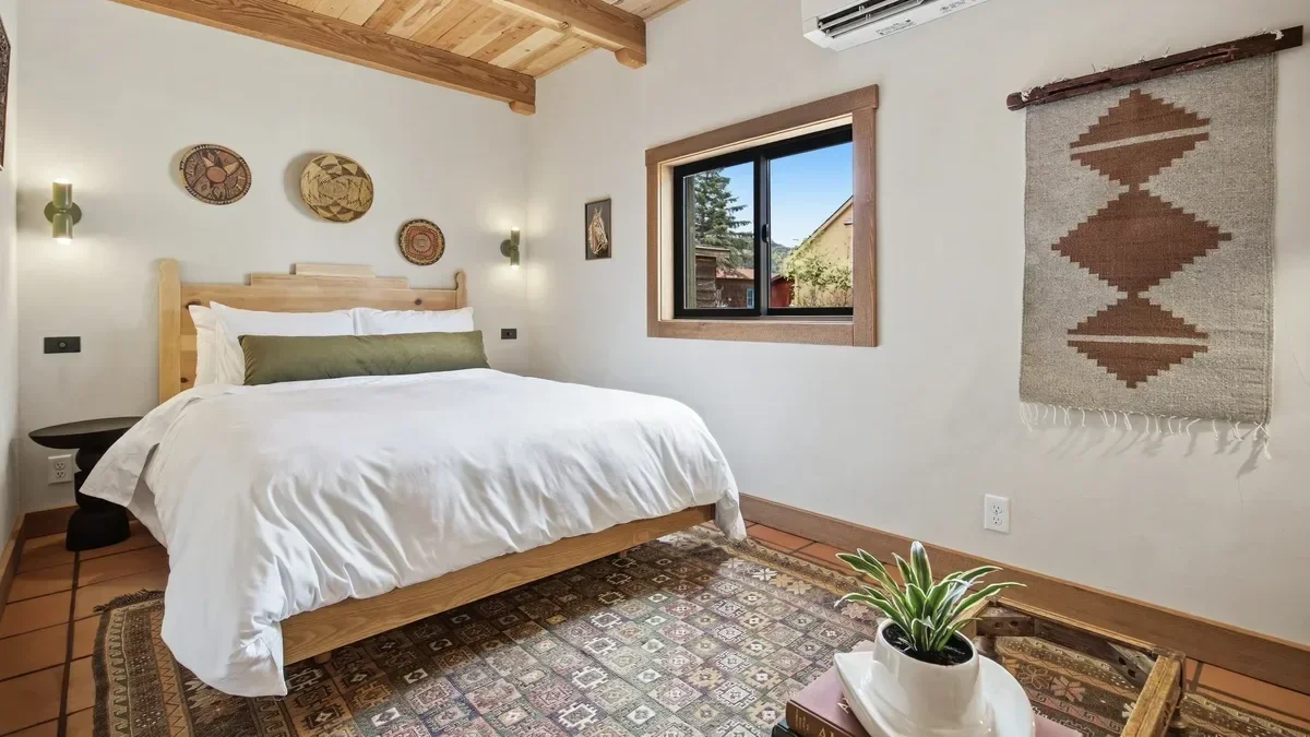 A cozy bedroom with a wooden ceiling and trim, a white bed with an olive green bolster pillow, wall-mounted lights, a window showing trees and a house outside, a decorative wall hanging, a potted plant on a small table, and a patterned rug on the flo