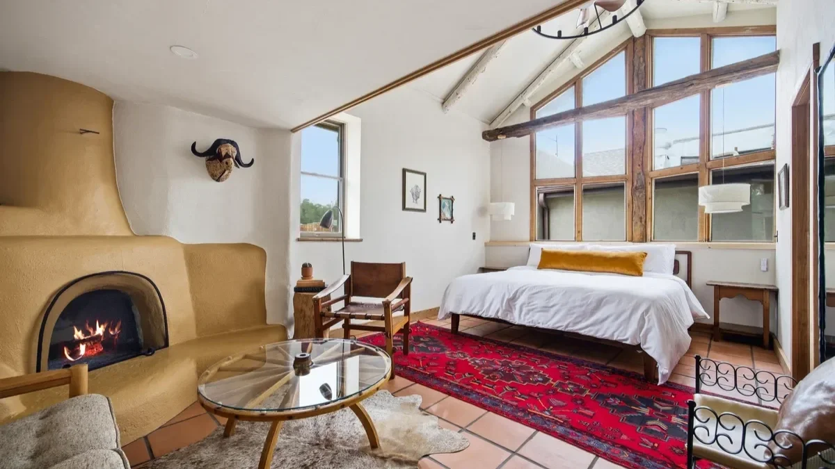 Cozy bedroom with white walls and a high ceiling with wooden beams, large windows, a white bed with a gold pillow, a red patterned rug, a fireplace, and various furniture pieces including chairs and side tables.