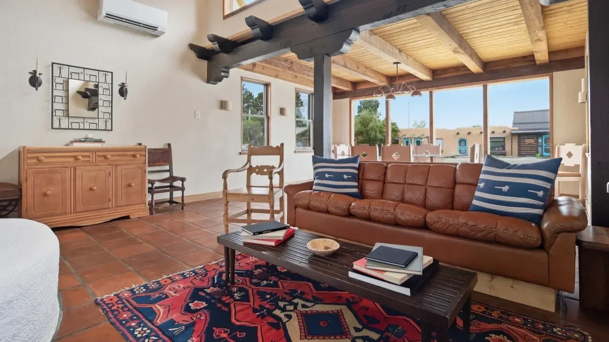 Living room with a brown leather sofa, wooden chairs, a coffee table with books and a bowl, a patterned rug, and large windows in a house with wooden ceiling beams.