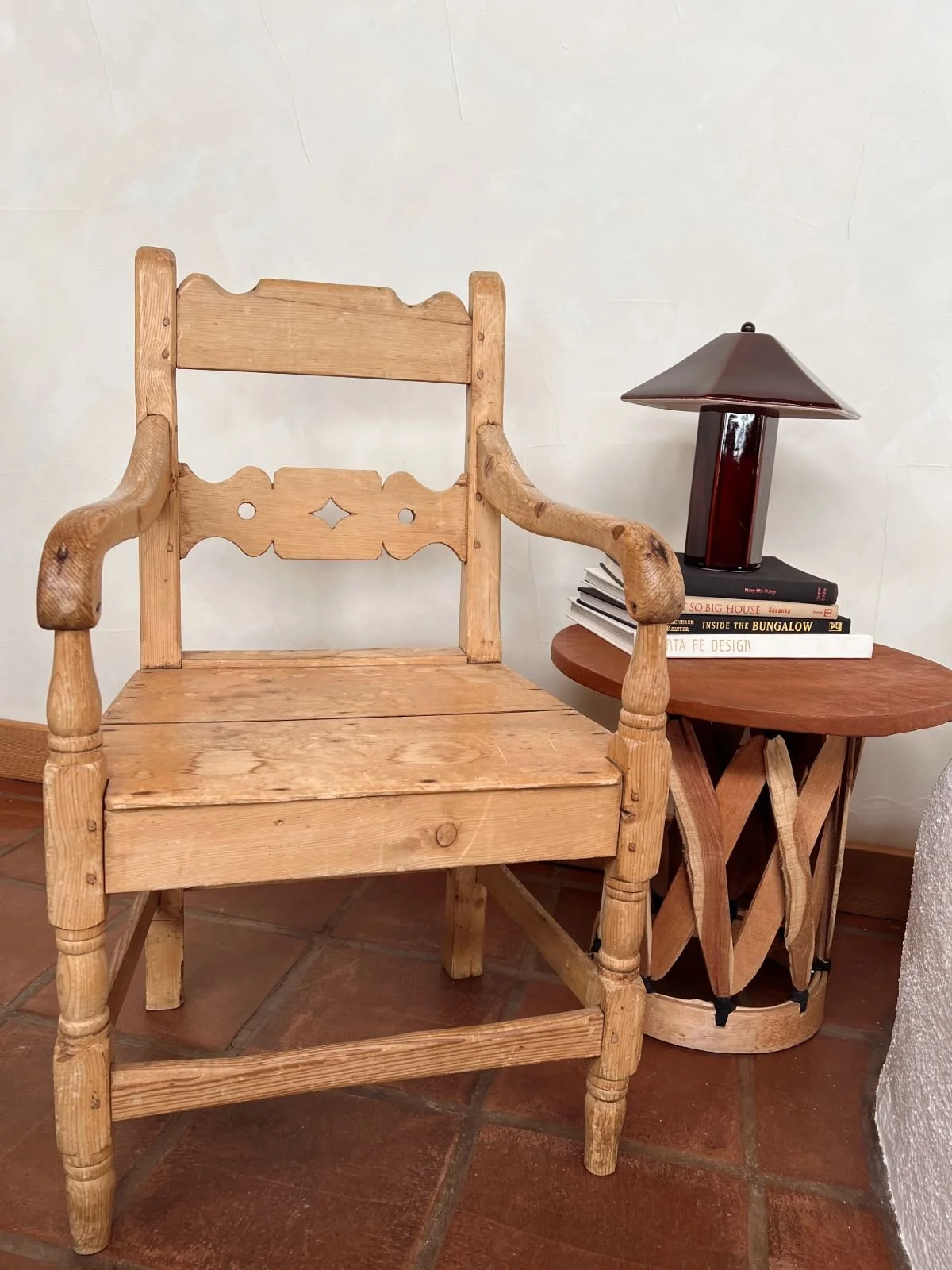 A wooden chair with a rustic finish next to a small side table holding a stack of books and a dark-colored lamp with a pyramid-shaped shade.