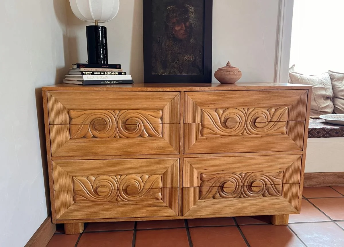 Wooden dresser with carved swirl patterns on its four drawers, topped with a lamp, a stack of books, a framed picture, and a small clay pot, in a room with tiled floor and beige walls.