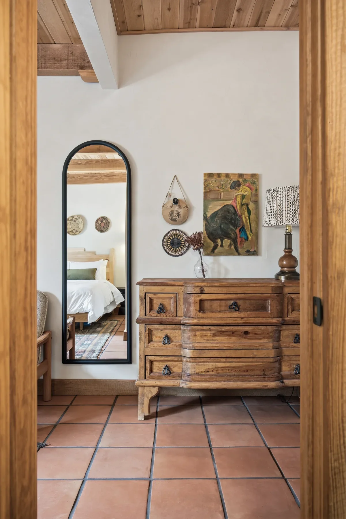 View through doorway showing a bedroom with a wooden bed, a mirror on the wall, and a wooden dresser with decorative items on top, including a lamp and a vase with dried flowers. The room has a wooden ceiling and terracotta tiled floor.