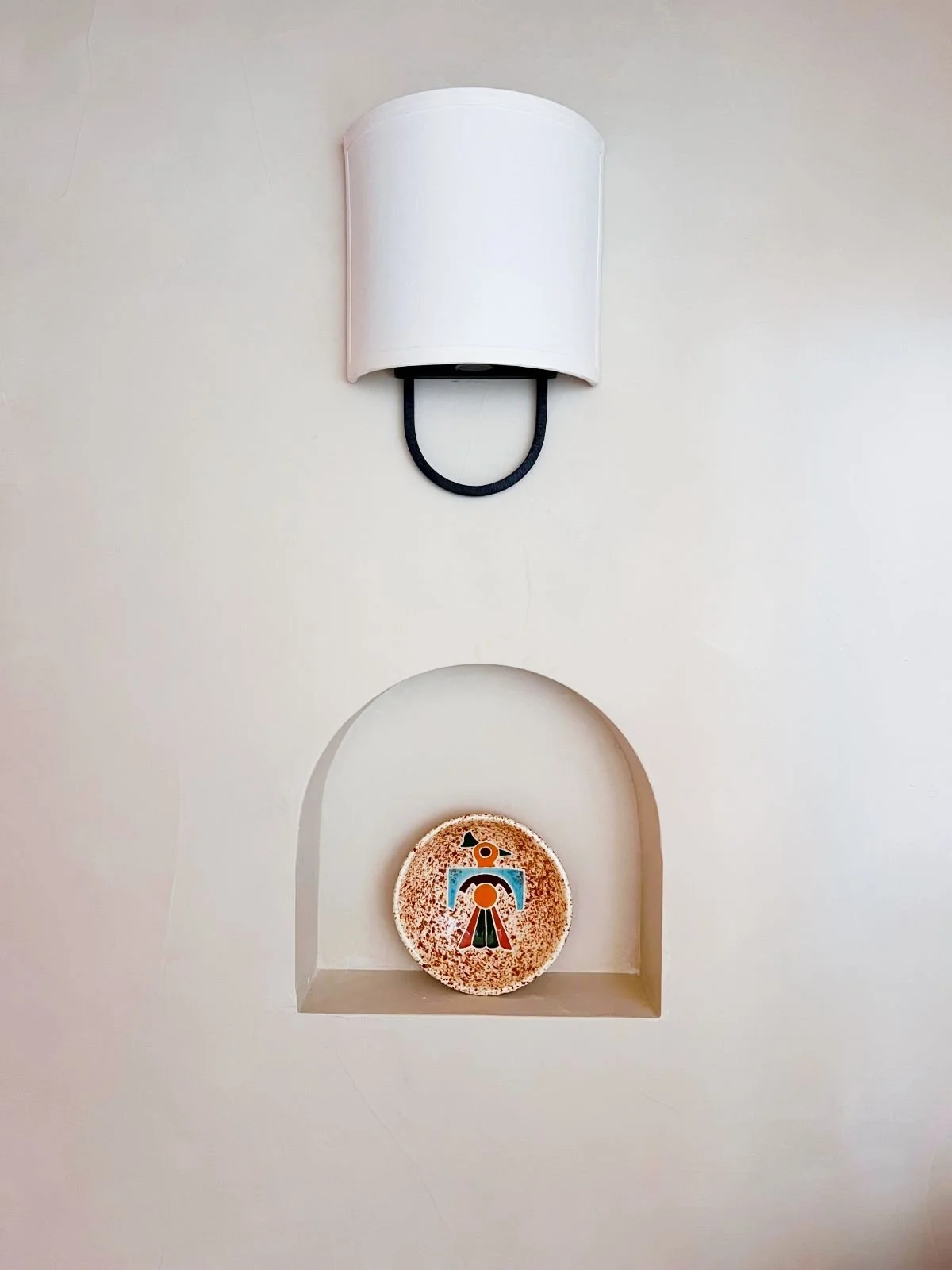 A white wall with a rounded white wall lamp above, and a decorative round plate with a stylized graphic of a clown or jester in the smaller arch below.