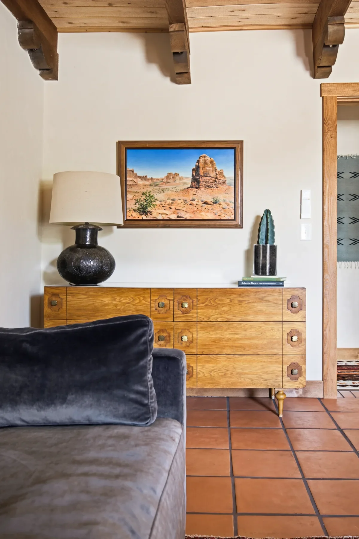 Interior of a living room featuring a wooden sideboard with square pattern drawer handles, a black ceramic table lamp with a beige lampshade, a framed desert landscape photograph, and a tall green cactus sculpture on a stack of books. The room has terracotta tile flooring, exposed wooden ceiling beams, and wooden doorframe on the right.