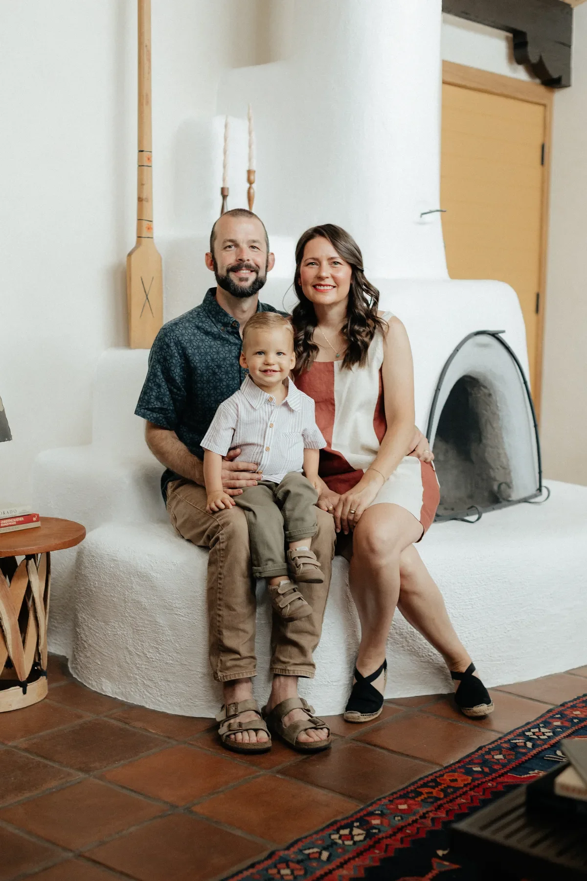 A family of three sitting on a white textured couch in a cozy living room, smiling at the camera. The father has a beard and is wearing a dark patterned shirt and khaki pants. The mother has long dark hair, wearing a sleeveless dress. Their young son, sitting on his father's lap, has short blond hair, wearing a striped shirt and khaki shorts.