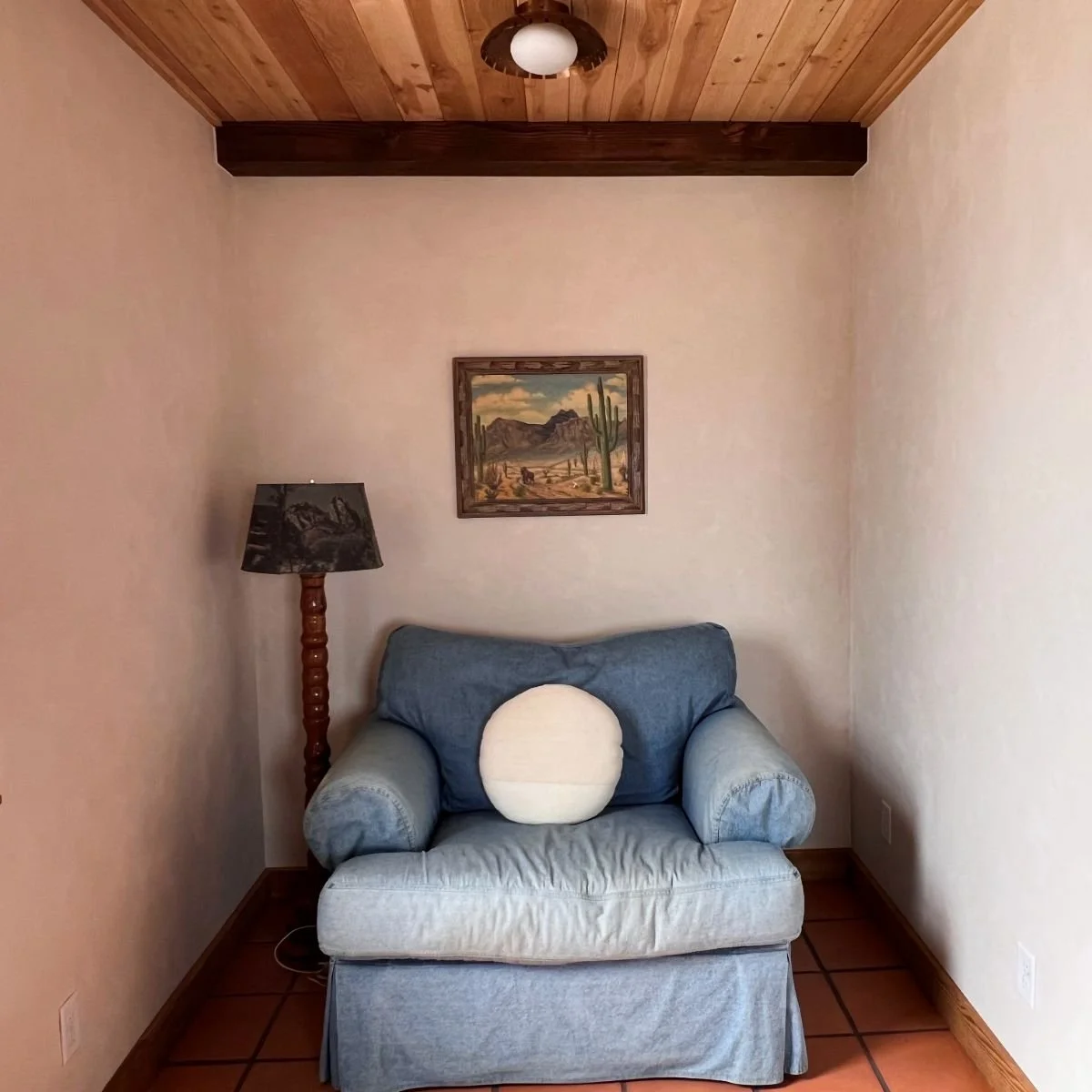 A cozy living room corner featuring a light blue armchair with a round white cushion, a tall wooden floor lamp with a black lampshade, a framed painting of a desert landscape with cacti on the wall, and a wooden ceiling with a ceiling light. The room has beige walls and a terracotta tile floor.
