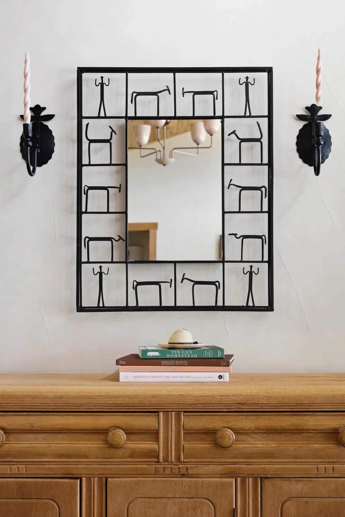 A wooden sideboard with a stack of books and a small ceramic object on top. Above it, a decorative mirror with a black metal frame featuring stick figure drawings of people and tables. Two black wall sconces with pink twisted candles are on either side of the mirror. A chandelier with multiple white shades is reflected in the mirror.
