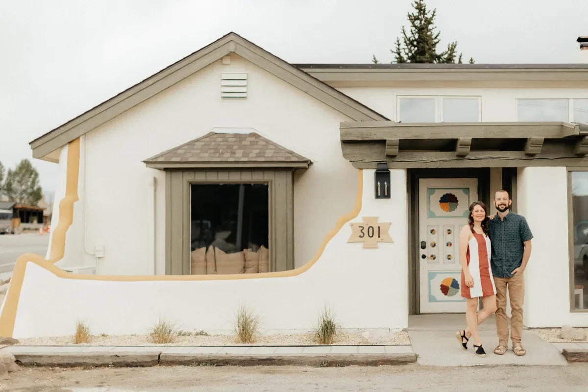 A couple standing in front of a house with an artistic exterior featuring a white wall with beige and green accents, a window, and a door decorated with colorful fan designs, with the house number 301 displayed next to the door.