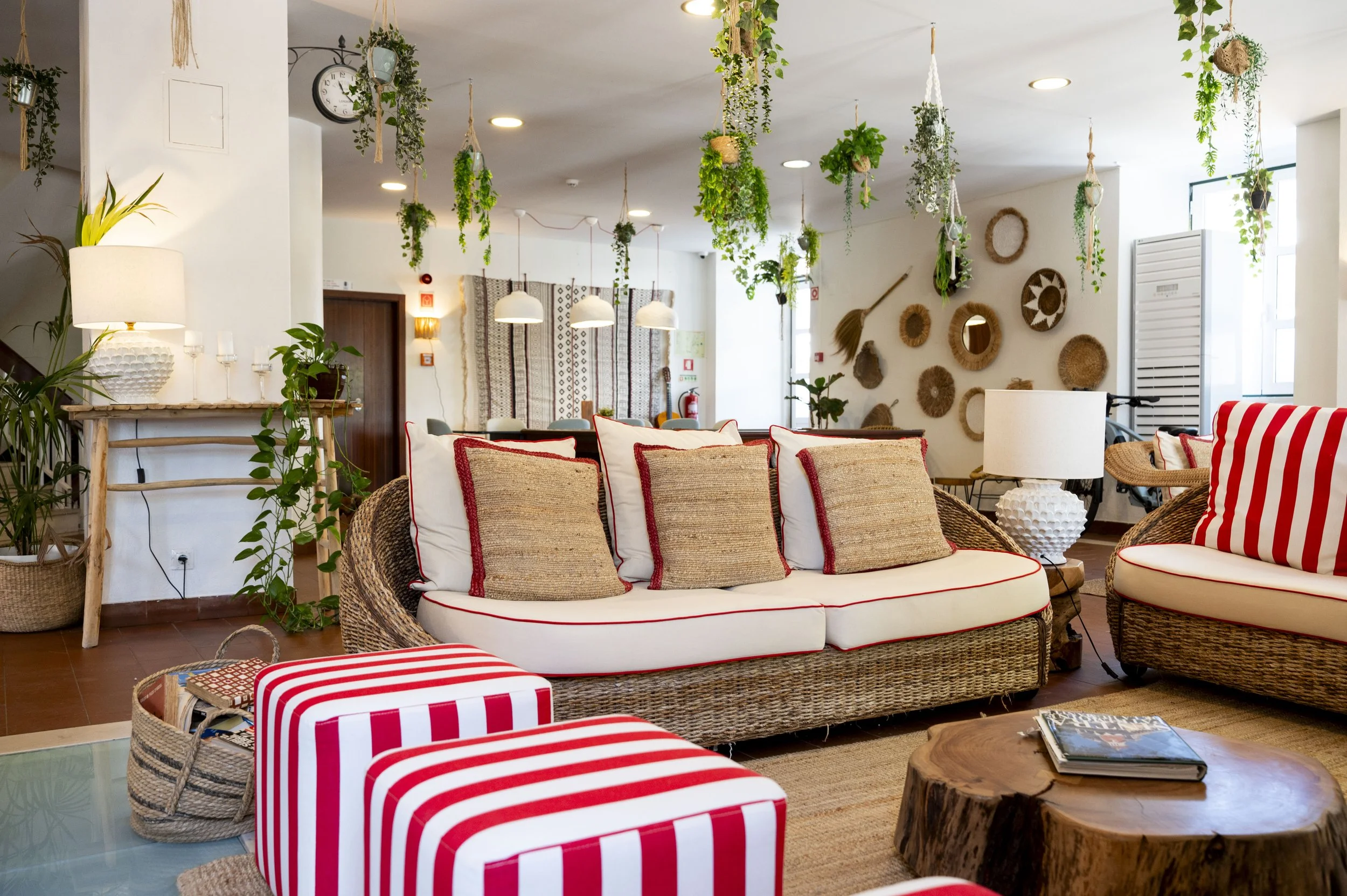 Sala de estar decorada com móveis de fibra natural e almofadas, com parede decorada com objetos de palha, plantas penduradas e luminárias suspensas, criando ambiente aconchegante e rústico.