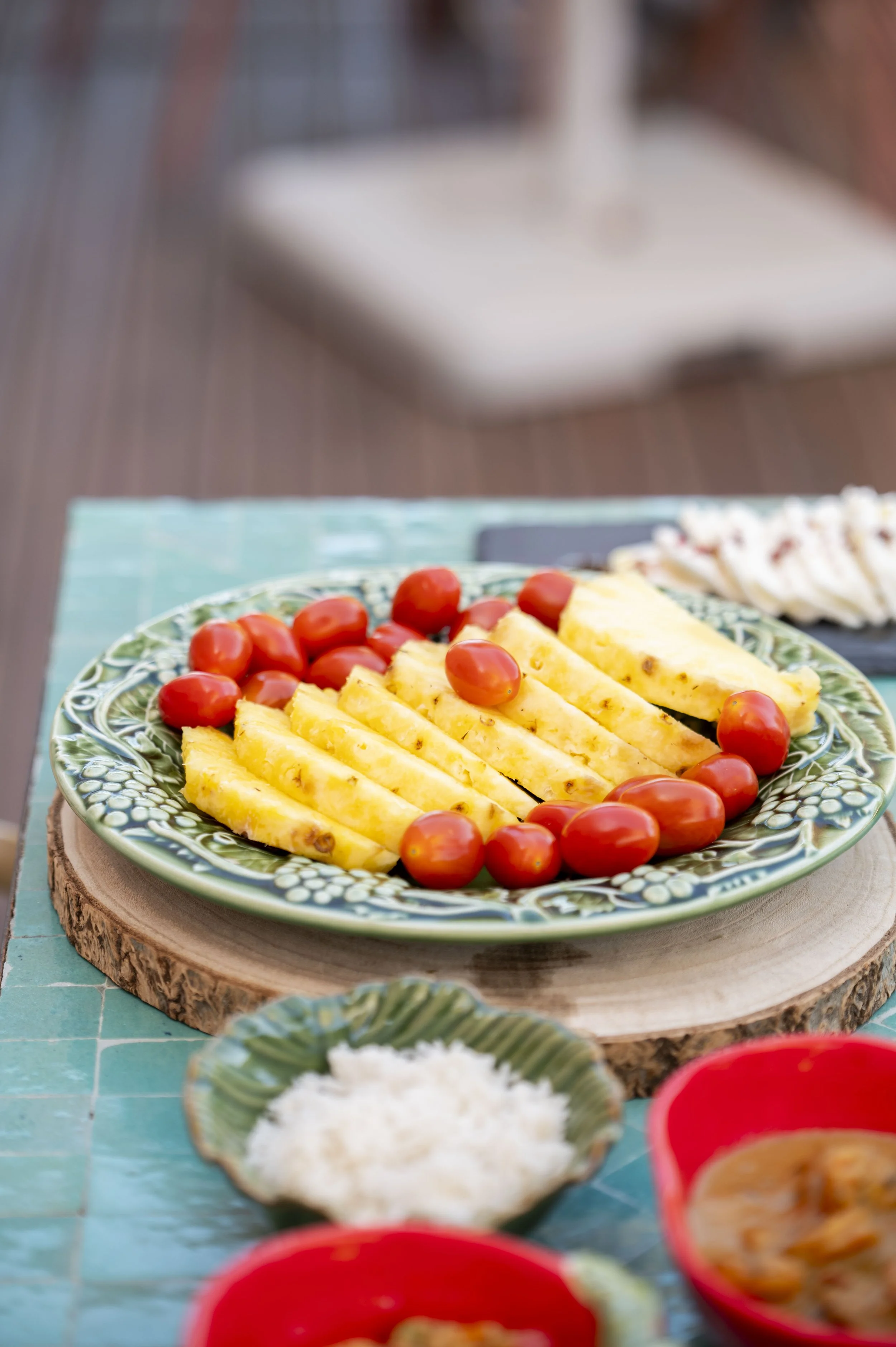 Prato com pedaços de abacaxi e tomates cereja, colocado sobre uma tábua de madeira. Ao fundo, uma bandeja de comida e tigelas vermelhas com alimentos variados.