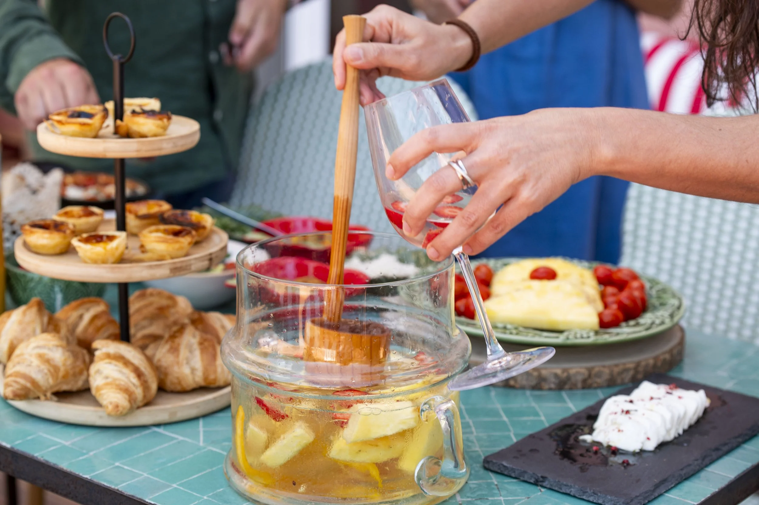 Pessoas em um evento ao ar livre servindo bebidas e comidas em uma mesa decorada com vários tipos de salgados, doces e frutas.